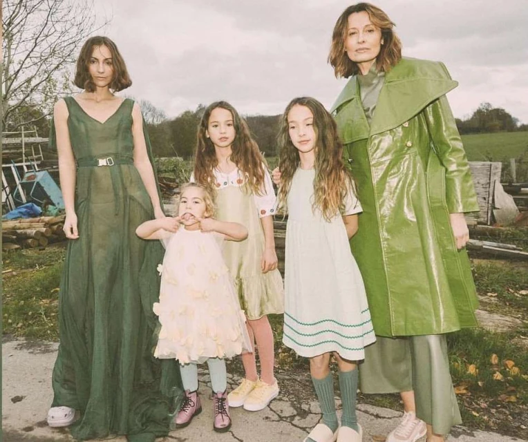 A group of five females in outdoor setting. Two adult women in dresses or coat, accompanied by three young girls in dresses.