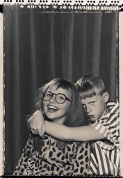 Black and white portrait of a smiling girl wearing round glasses and a boy in a striped shirt embracing her by photographer Ian Boddy.