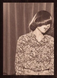 Monochrome portrait of a person with heavy bangs wearing a floral shirt against a draped curtain background