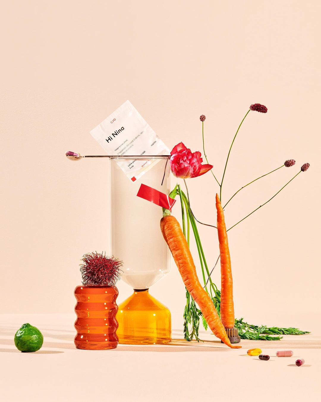 Still life featuring carrots, flowers, glass vases, lime, and supplements against a peach background in a visually striking composition.