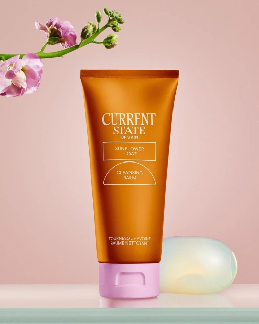 A skincare product tube, Current State of Skin cleansing balm, stands with a soft floral accent and soap, evoking beauty and self-care.