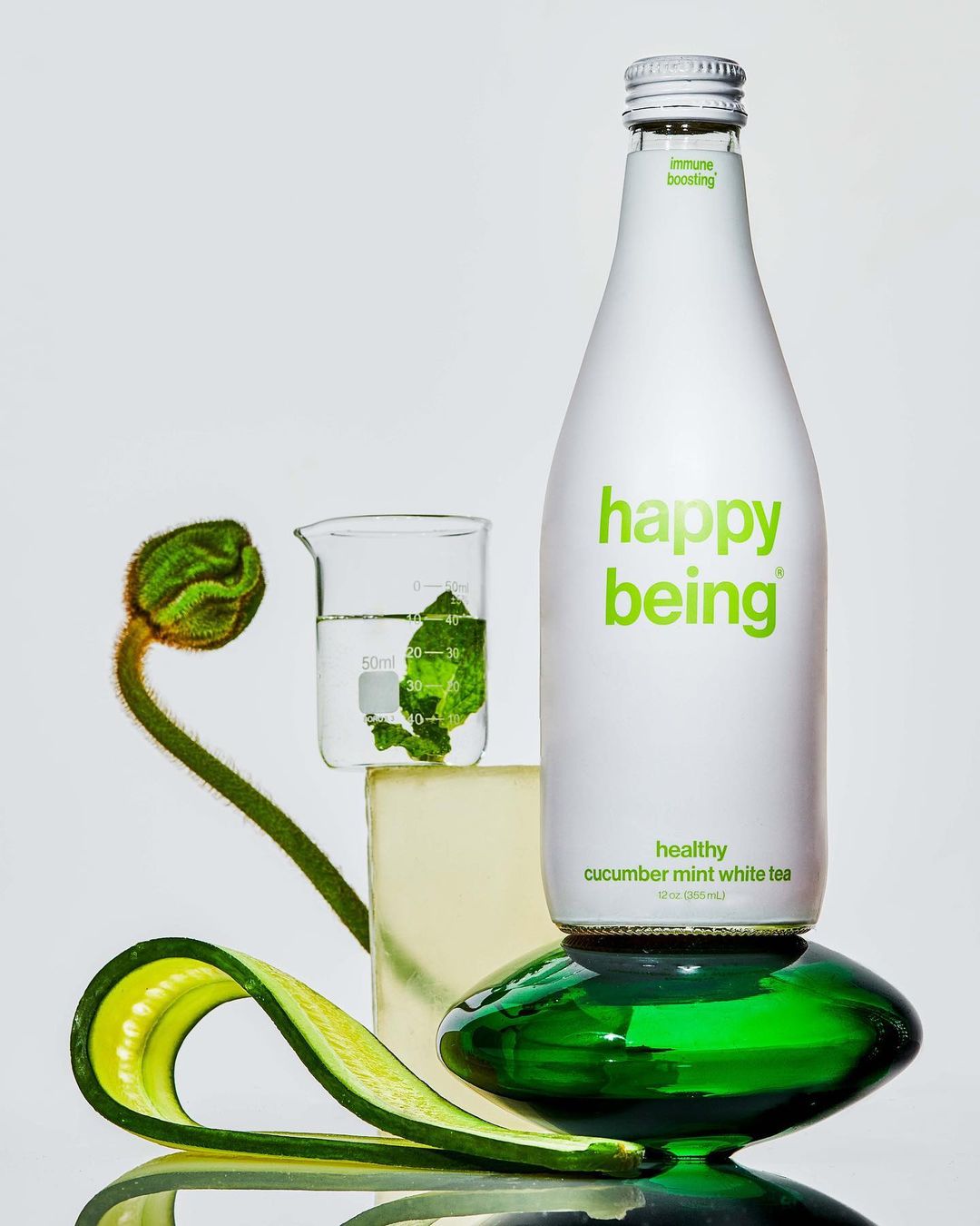 Still life of Happy Being cucumber mint white tea bottle on a green glass pedestal, with cucumber, flower bud, and beaker.