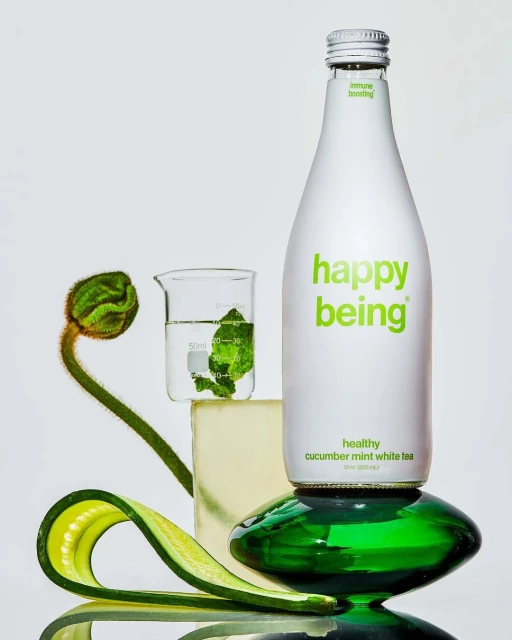 Still life of Happy Being cucumber mint white tea bottle on a green glass pedestal, with cucumber, flower bud, and beaker.