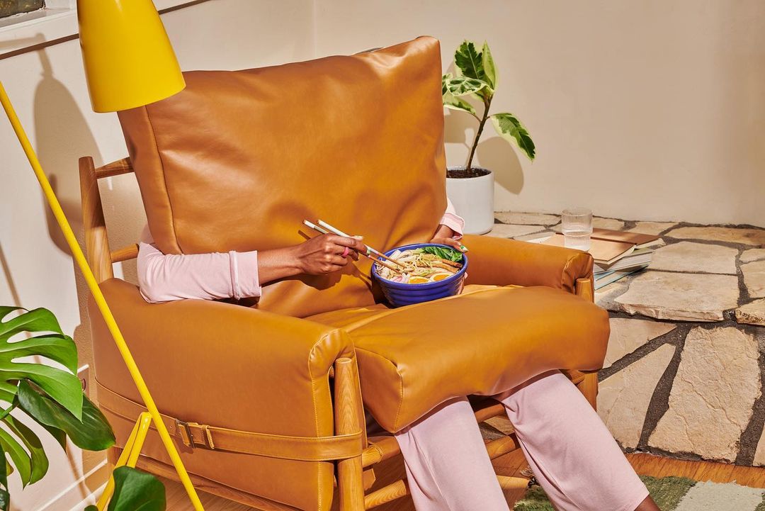 Person sitting in a brown leather chair eating noodles from a blue bowl with chopsticks indoors with a lamp and green plant.