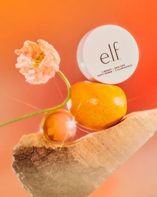 Beauty product shot: white ELF cosmetic container, orange, flower, and orb on a rock against an orange gradient background.