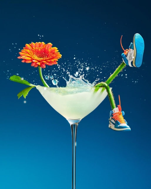 Still life shot of a gerbera daisy, sneakers, and cocktail in a martini glass against a blue background. Whimsical, floral, and refreshing.