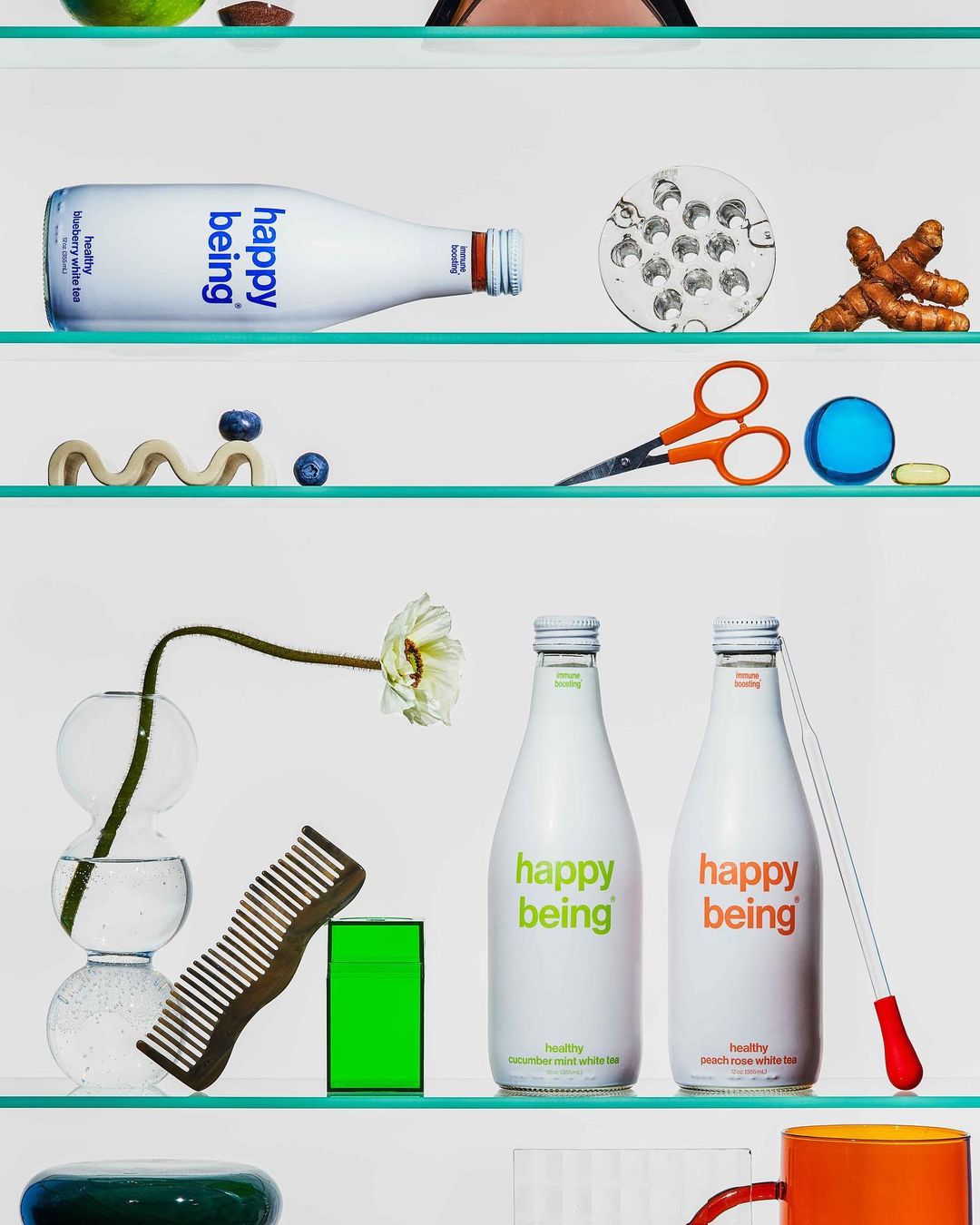 Still life on glass shelves featuring two bottles of Happy Being brand drink mix, a hair comb, a flower in a vase, and other items.