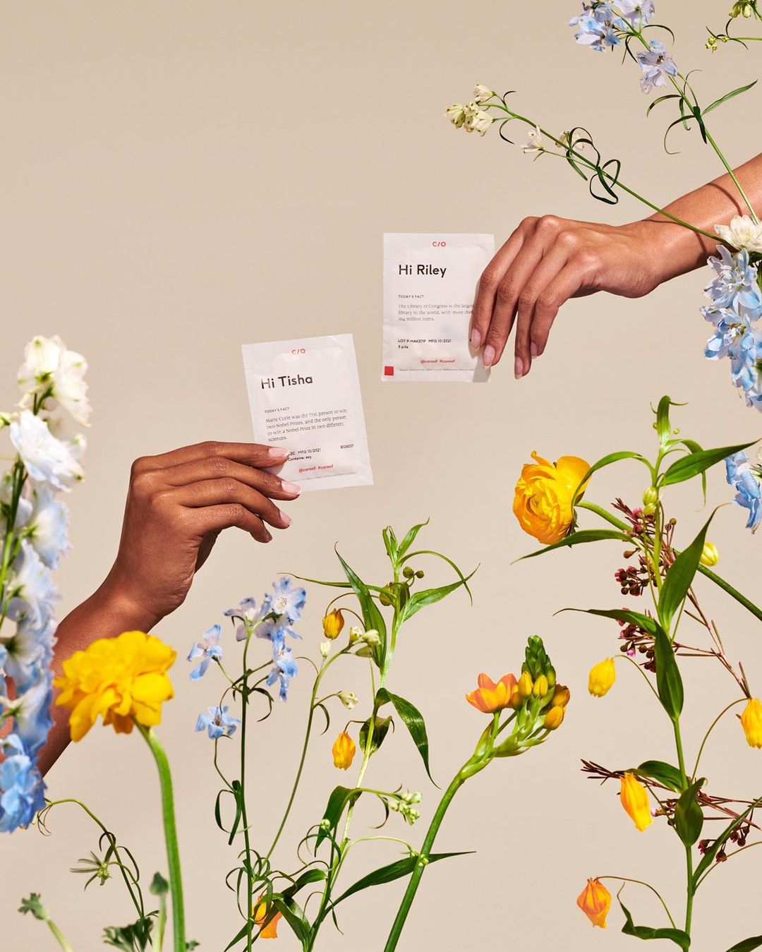 Two hands hold cards surrounded by vibrant spring flowers. The cards and flowers are set against a solid light beige background.