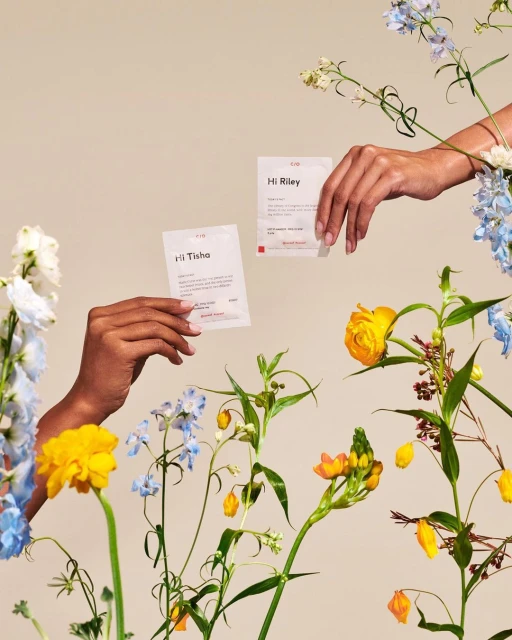 Two hands hold cards surrounded by vibrant spring flowers. The cards and flowers are set against a solid light beige background.