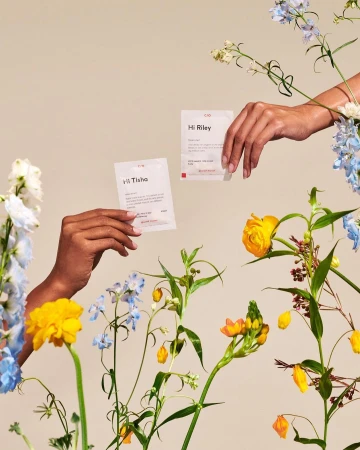 Two hands hold cards surrounded by vibrant spring flowers. The cards and flowers are set against a solid light beige background.