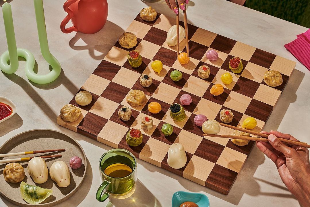 Overhead shot of a checkerboard game with colorful dumplings as pieces, placed indoors, with two people holding chopsticks, table.