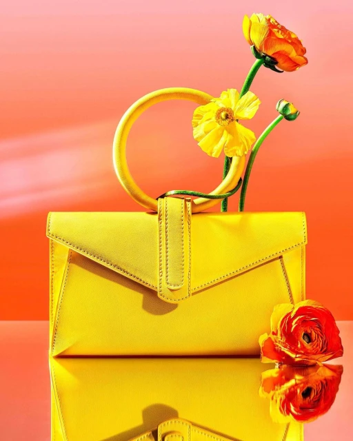 Yellow clutch bag with flowers on a reflective surface, set against an orange gradient backdrop, showcasing a vibrant and stylish composition.