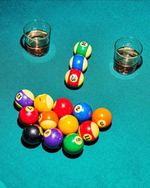 Overhead shot on a billiard table featuring billiard balls racked, two glasses of amber liquid, and green felt.