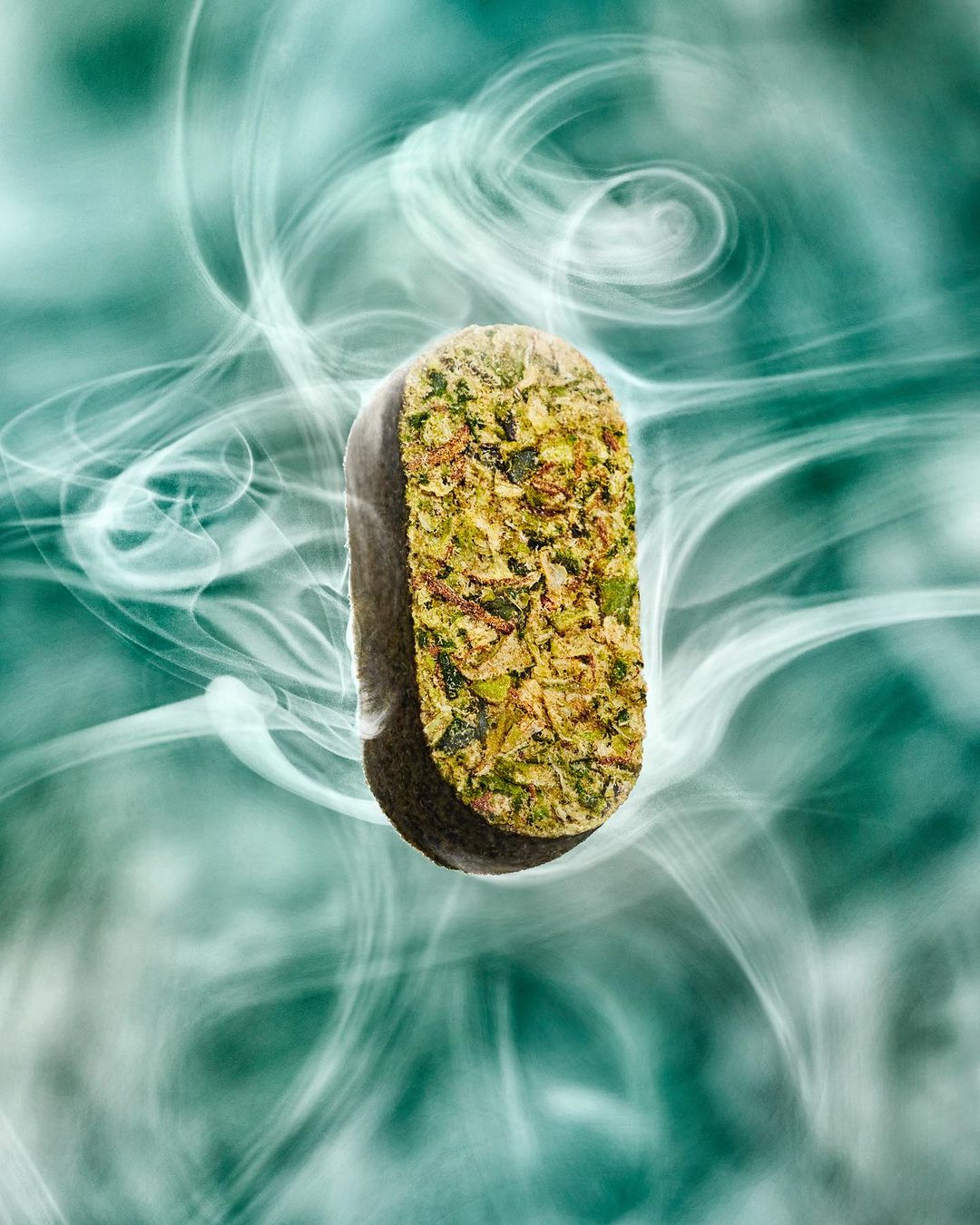 Abstract still life of an elongated capsule form featuring mottled green textures against an ethereal smoke and gradient teal background.