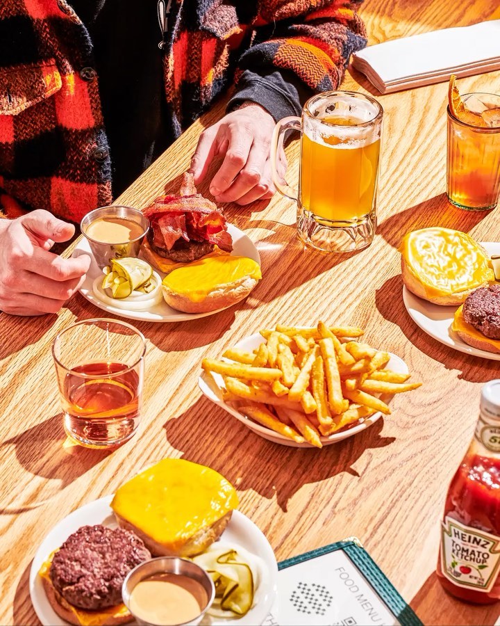 Overhead shot of burgers with fries and beverages on a wooden table, one person visible, indoor dining setting, warm lighting.