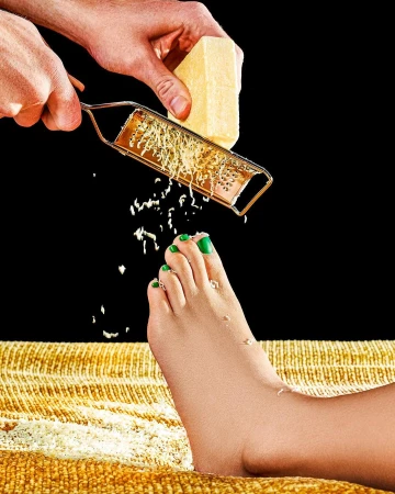 Person grating cheese onto a human foot with green nail polish, resting on a gold textured surface against a black background.