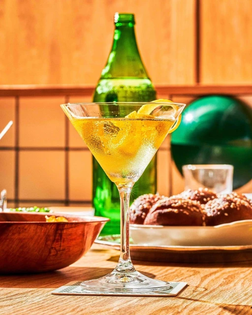 Close-up shot of a martini glass filled with a yellow cocktail and a lemon twist, set on a wooden table with a green bottle behind.