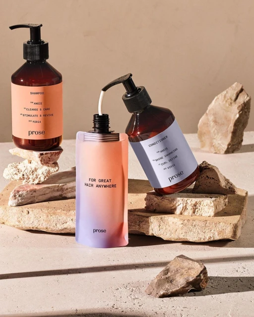 Three Prose brand hair product bottles in amber and gradient peach colors sit on sandstone slabs against a neutral backdrop in a studio shot.