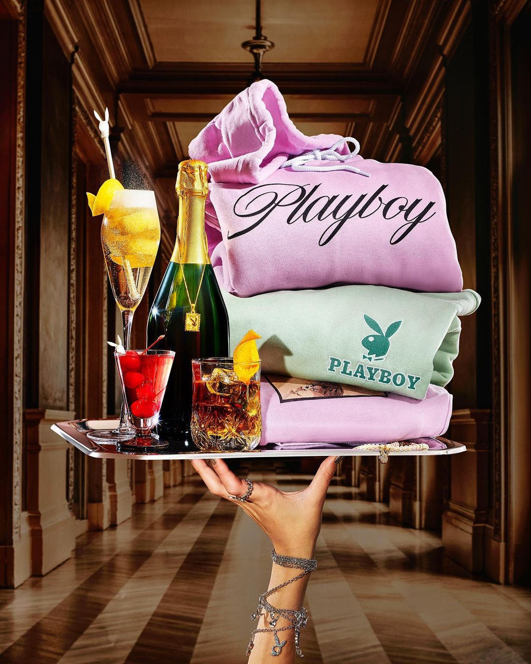 Hand holding a tray with drinks, champagne, and folded Playboy hoodies with a luxury hallway background.