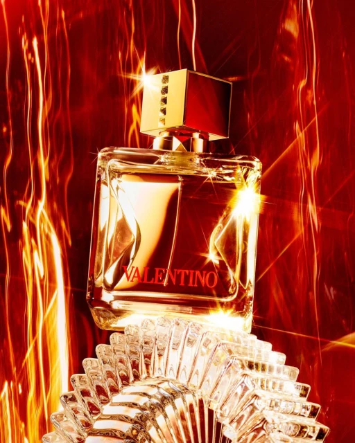 Valentino perfume bottle on textured clear glass pedestal, red and gold accents, abstract light trails, beauty and fashion product.