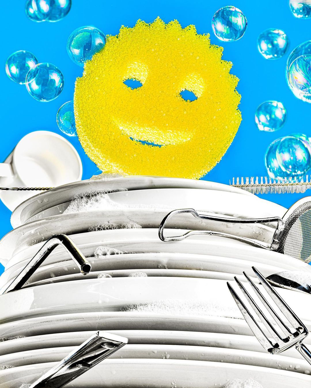 Stacked clean dishes with a yellow smiling sponge, soap bubbles, and dishwashing utensils against a bright blue background. Food & Beverage.