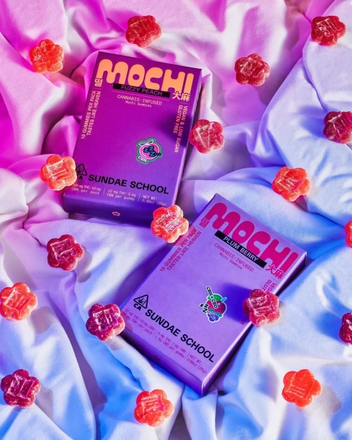 Two boxes of cannabis-infused gummies by Sundae School are on a white crumpled fabric surface surrounded by additional gummy candies.