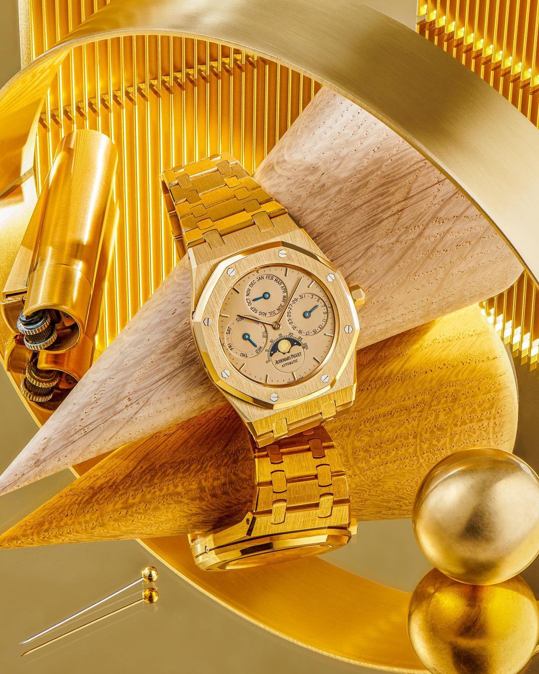 Elevated view of a luxurious gold Audemars Piguet watch on a wood prop surrounded by golden metal accents, set against a golden background.