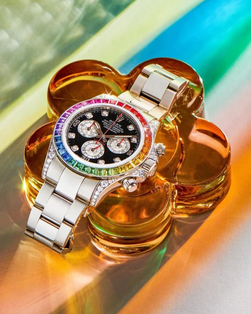 Close-up of a luxury wristwatch with a rainbow gemstone bezel, resting on a clear, flower-shaped platform, with rainbow reflections in the background.