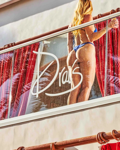 Blonde woman in blue bikini stands on a balcony overlooking a pool, with 'Drai's' written on the glass partition.