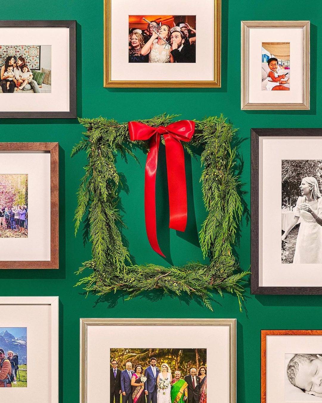 Framed family photos are displayed on a green wall, surrounding a square evergreen wreath with a large red ribbon, capturing family memories.