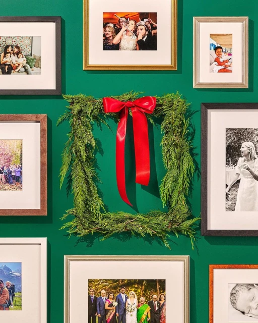 Framed family photos are displayed on a green wall, surrounding a square evergreen wreath with a large red ribbon, capturing family memories.