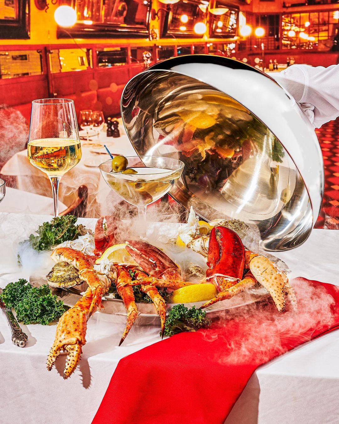 Seafood platter with lobster, oysters, and champagne on a table, presented with a silver cloche in a restaurant setting.