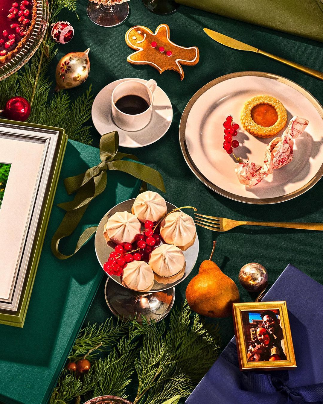 Festive holiday table setting with coffee, cookies, dessert, ornaments, and decorations on a dark green tablecloth for a Christmas celebration.