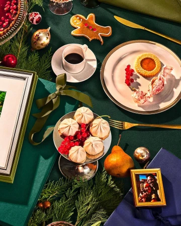 Festive holiday table setting with coffee, cookies, dessert, ornaments, and decorations on a dark green tablecloth for a Christmas celebration.