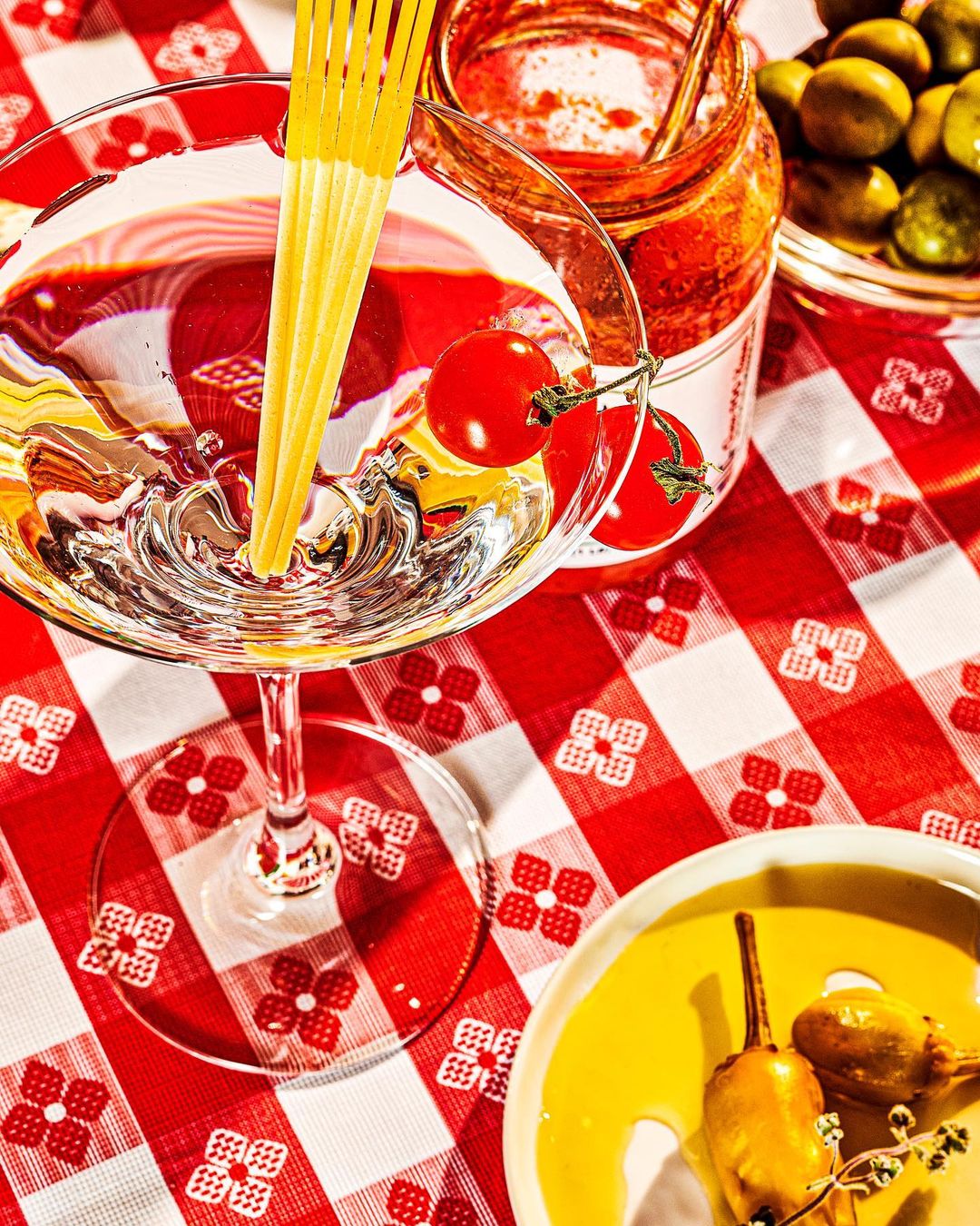 Overhead shot of a martini glass filled with pasta and cherry tomato garnish, set on a red and white checkered tablecloth with pickled peppers.