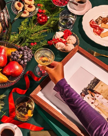 Overhead shot of table setting with holiday treats, fruit, drinks, and a framed photo being held by a person in a purple velvet shirt.
