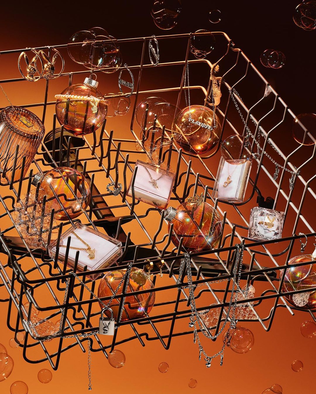 Still life of jewelry pieces and perfume bottles artfully arranged inside a dishwasher rack against an orange gradient background.
