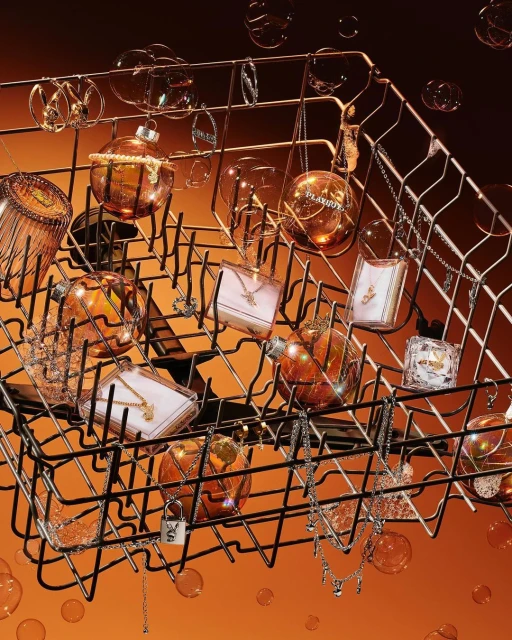 Still life of jewelry pieces and perfume bottles artfully arranged inside a dishwasher rack against an orange gradient background.