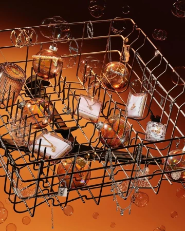 Still life of jewelry pieces and perfume bottles artfully arranged inside a dishwasher rack against an orange gradient background.