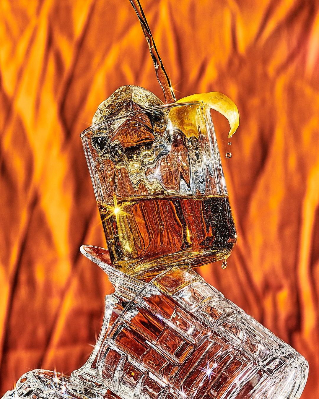 Close-up of crystal glasses stacked, one filled with whiskey and ice, a lemon peel, and a stream of liquid against an orange background.