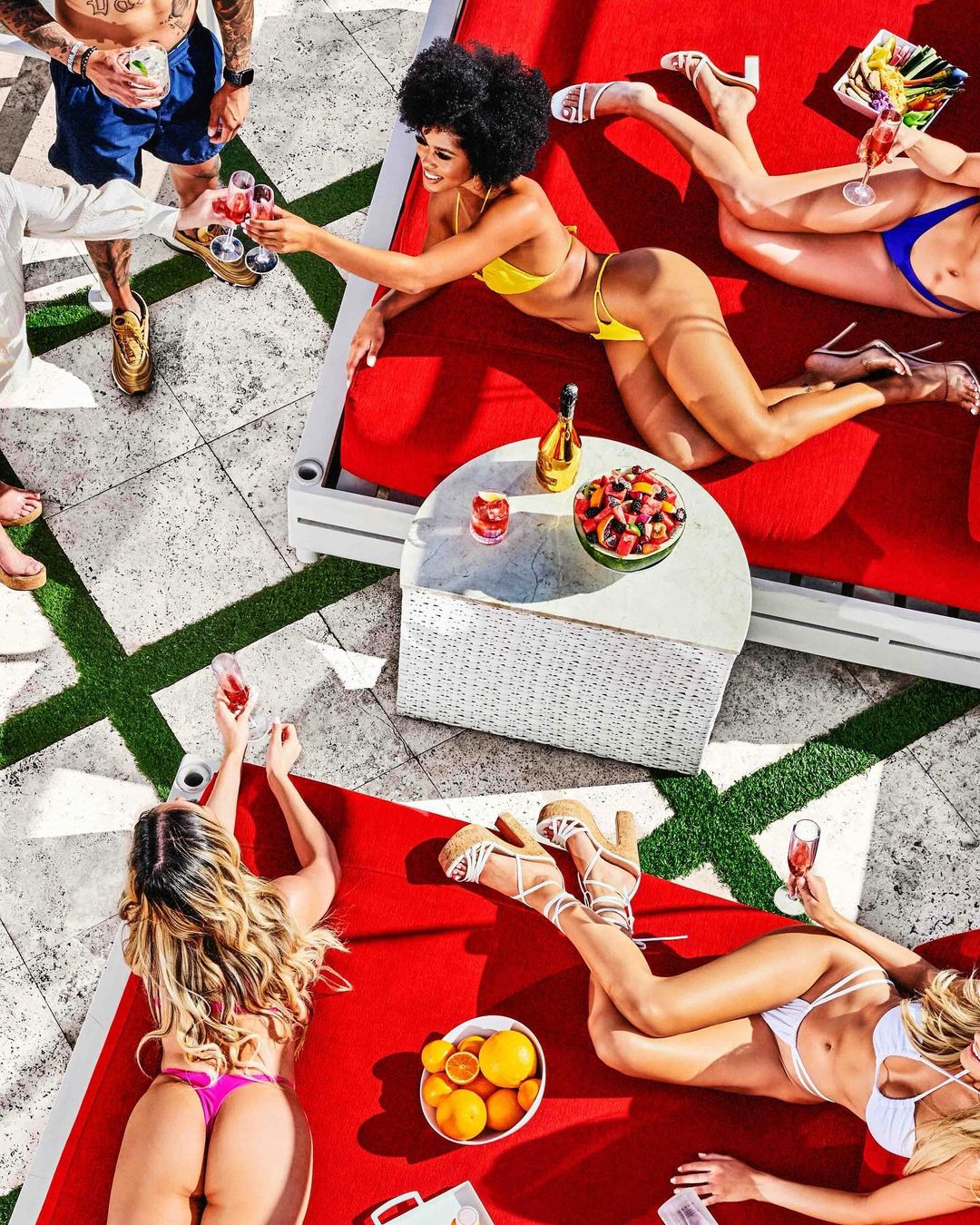 Overhead shot of women in swimsuits lounging on red lounge chairs by a pool, enjoying drinks and fruit in an outdoor setting.