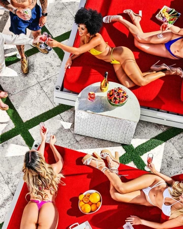 Overhead shot of women in swimsuits lounging on red lounge chairs by a pool, enjoying drinks and fruit in an outdoor setting.