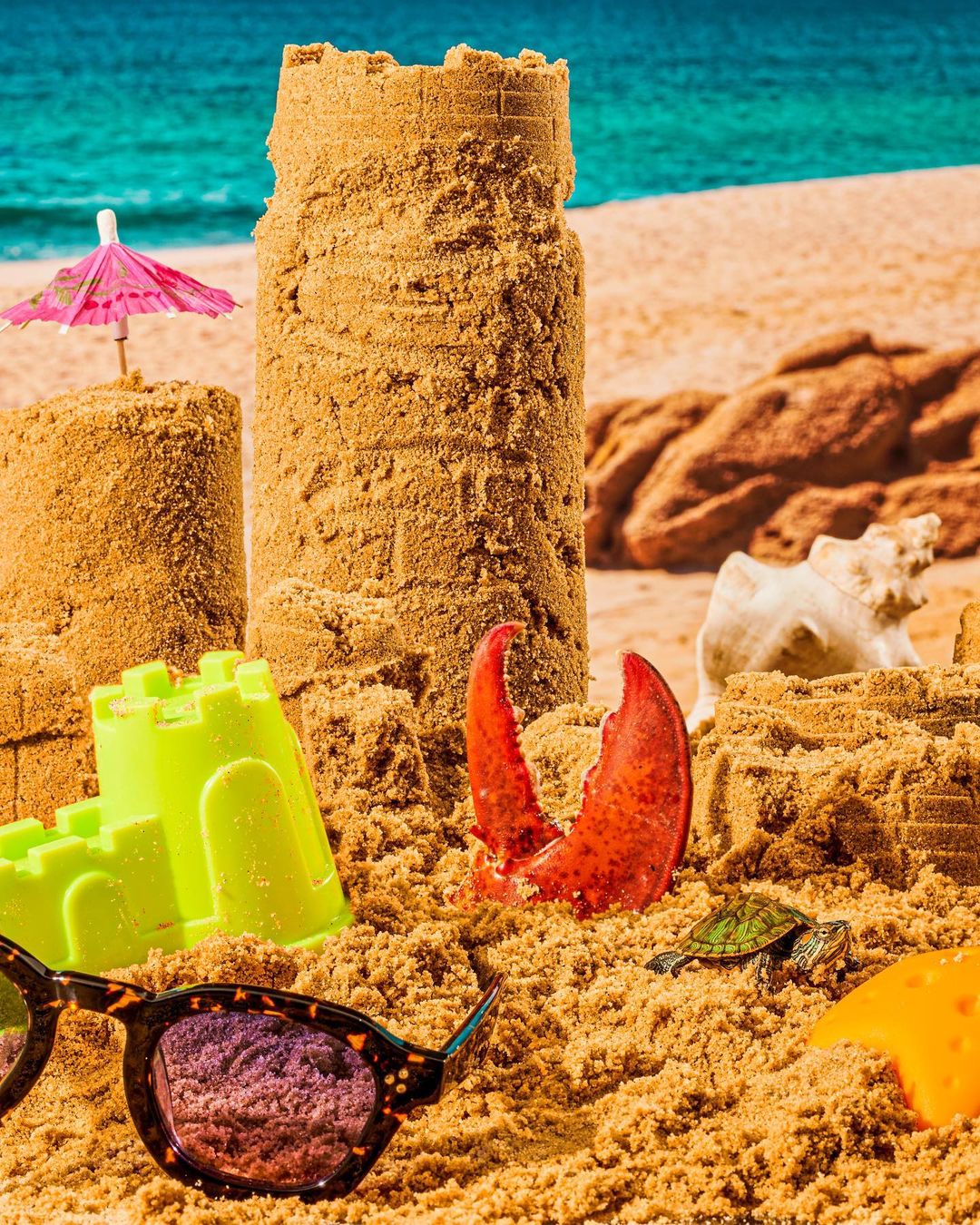 Beach scene with large sandcastles, sunglasses, toy turtle, and lobster claw, set against ocean backdrop on bright sunny day.