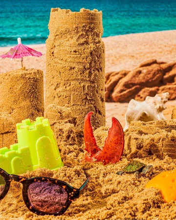 Beach scene with large sandcastles, sunglasses, toy turtle, and lobster claw, set against ocean backdrop on bright sunny day.