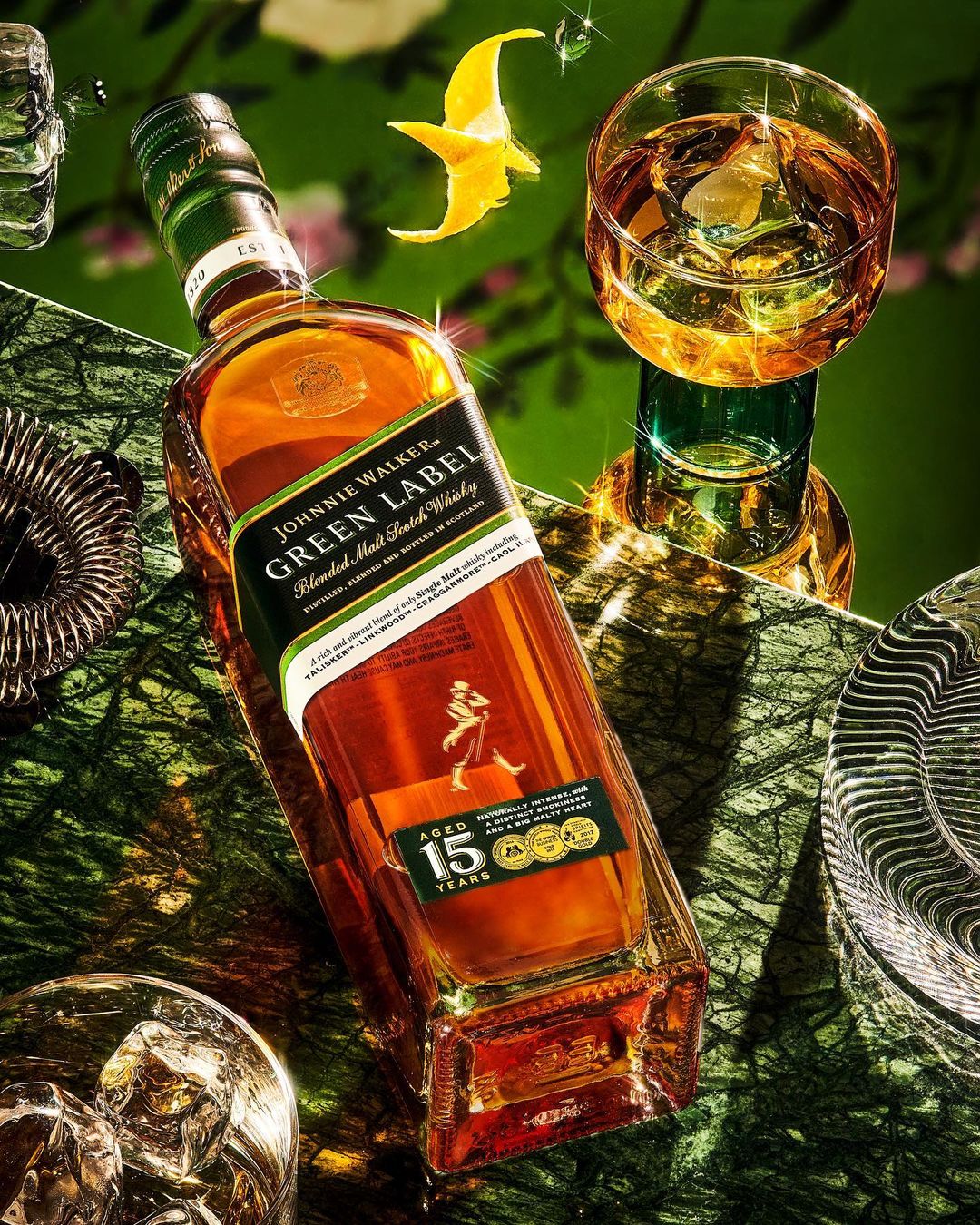 Johnnie Walker Green Label bottle on a marble surface with a glass of whiskey, citrus peel, and bar tools, showcasing luxury.