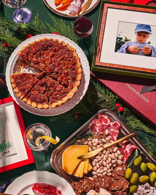 Overhead shot of pecan pie, charcuterie board, holiday greenery, and framed picture on a festive green surface, celebrating food.