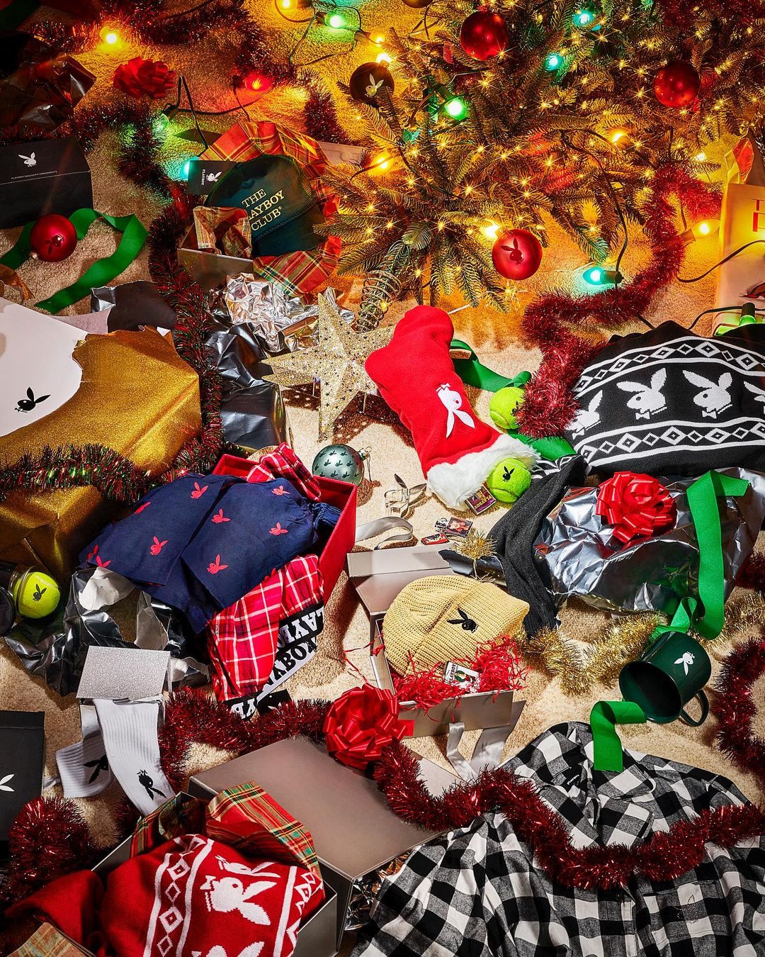 Playboy-themed holiday display with gifts, including apparel, under twinkling Christmas lights and a decorated tree. Presents are open.