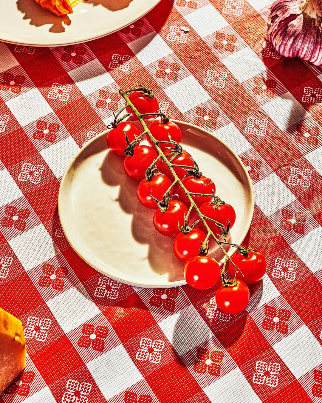 Close-up of a plate with a vine of red cherry tomatoes resting on a red and white checkered tablecloth. Overhead shot, bright lighting.
