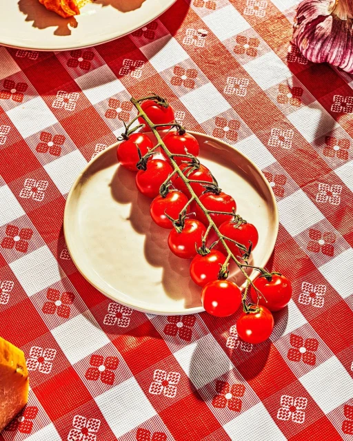 Close-up of a plate with a vine of red cherry tomatoes resting on a red and white checkered tablecloth. Overhead shot, bright lighting.