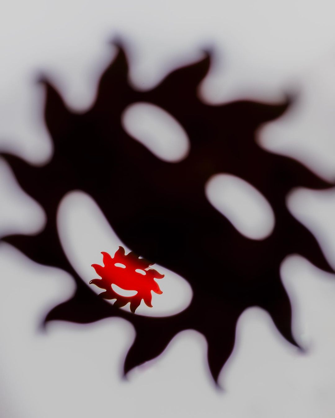 Abstract art featuring a large dark serrated shape with a smaller red serrated shape visible through an opening on a white backdrop.